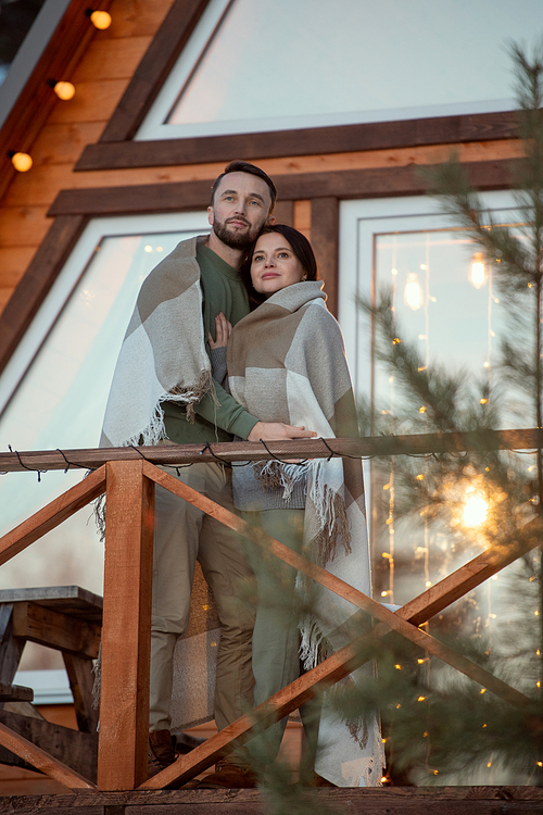 Young affectionate couple wrapped in plaid standing close to one another by new large country house and looking at sunrise in the morning