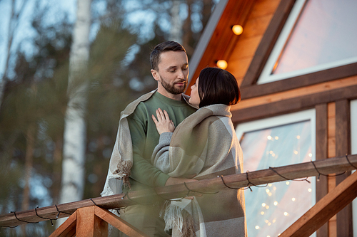 Happy young affectionate couple wrapped in plaid looking at one another while standing by new large country house in natural environment