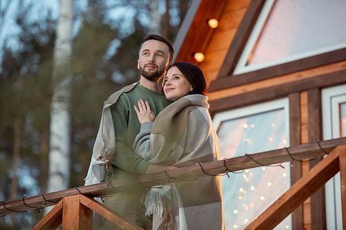 Young affectionate couple wrapped in plaid standing against country house on background of trees in the forest and looking at sunrise