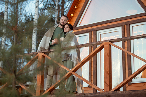 Young affectionate man and woman wrapped in plaid standing by their country house on background of trees and looking at sunrise