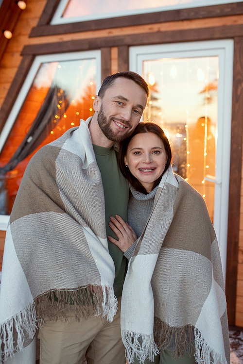 Young affectionate couple wrapped in plaid standing in front of camera against window of their new country house and looking at you