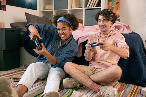 Happy teens with joysticks playing video game on bedroom floor by bed