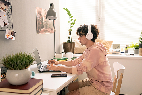 Young man with headphones sitting by desk and networking in front of laptop