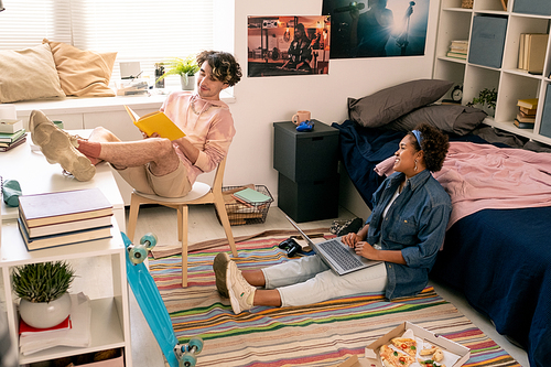 Male teenager reading book while his African girlfriend using laptop in bedroom