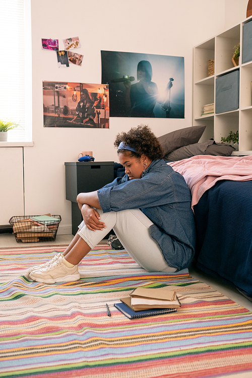 Troubled and lonely girl sitting on the floor by her bed and embracing her legs