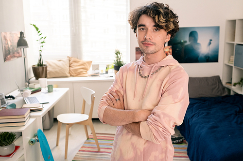 Cross-armed teenager in pink hoodie looking at camera in his bedroom