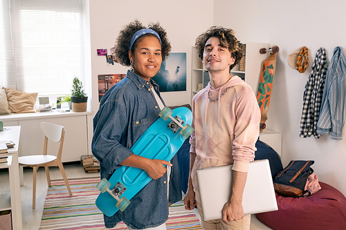 Happy intercultural teenagers with skateboard and laptop standing in bedroom