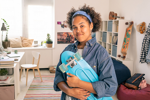 Pretty African female teenager with blue skateboard standing in her bedroom