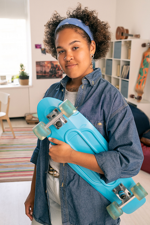 Happy African teenage girl in casualwear holding blue skateboard