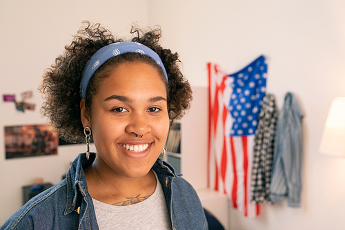 Happy African teenager in casualwear standing on background of American flag in her room