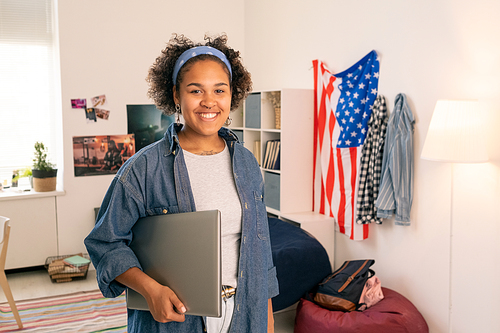 Cheerful American female teenager with laptop standing in her bedroom