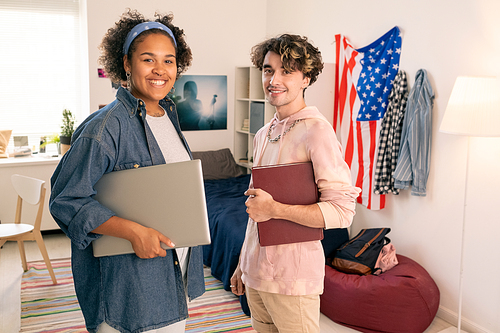 Two cheerful American teenagers in casualwear standing against national flag in domestic room