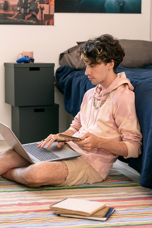 Young casual man with notepad sitting on the floor by bed and using laptop