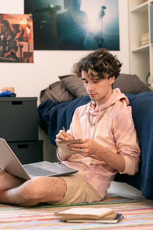 Teenage guy making notes in notepad while sitting on the floor by his bed