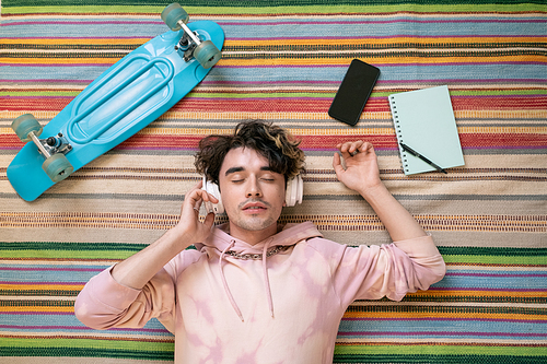 Restful guy in headphones and pink hoodie listening to music on the floor among skateboard, notepad and smartphone