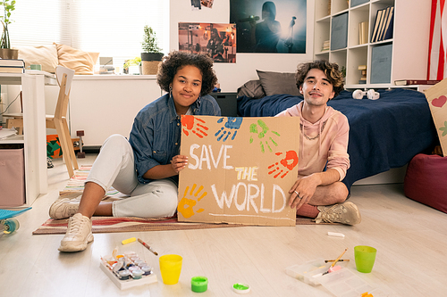 Happy teens showing handmade placard while sitting on the floor in bedroom