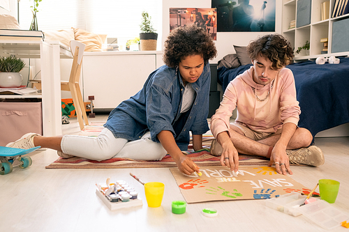 Intercultural teenage couple drawing placard on piece of cardboard while sitting on the floor