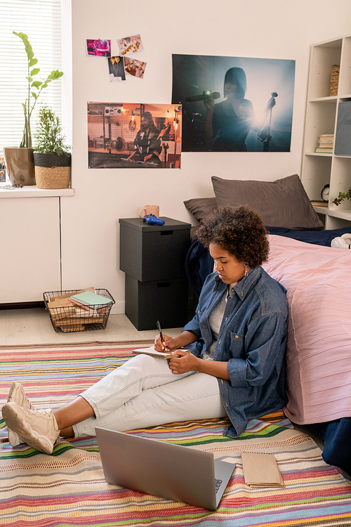 Adolescent girl in casualwear carrying out home assignment while sitting on the floor of bedroom