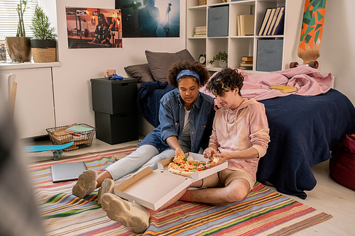 Hungry teenagers sitting on the floor by bed and having appetizing pizza