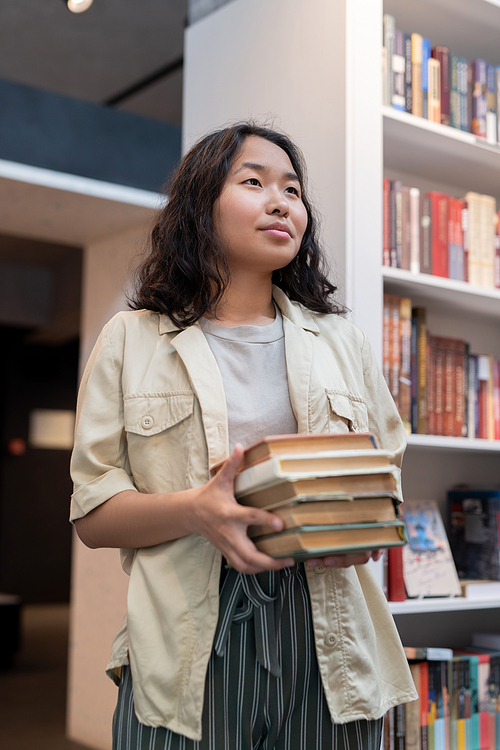 Contemporary student with stack of books standing against shelf in library