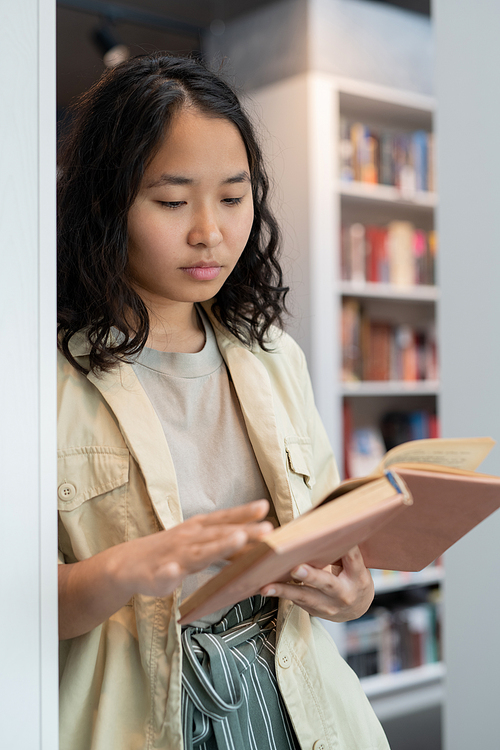 Clever student of college reading book while standing by bookshelf in library