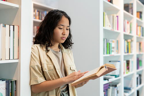 Serious female student with open book reading text by shelf in library at break