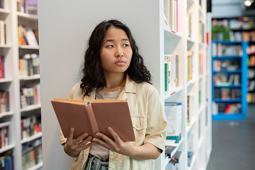 Young Asian woman holding open book while standing against bookshelf in library