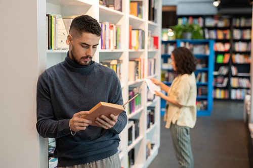 Serious young man reading book against bookshelf with brunette girl on background