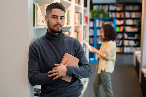 Bearded young man with book standing in front of camera against female choosing literature