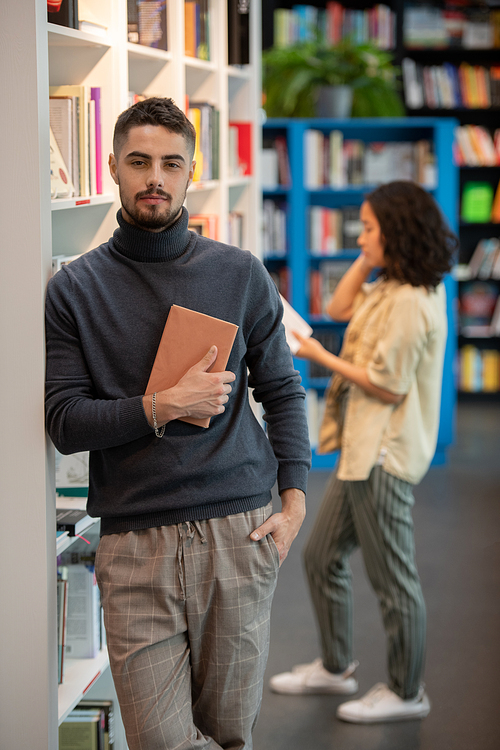 Young student in casualwear holding book by his chest with girl on background