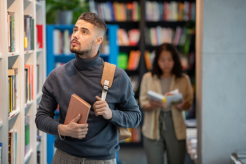 Pensive young man with book and backpack looking at bookshelf in college library