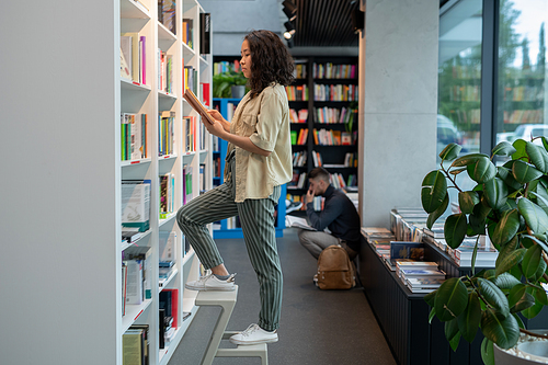 Pretty Asian woman standing on stepladder while choosing books in library