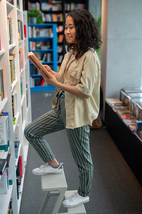 Cute Asian female student reading book by bookshelf in large library