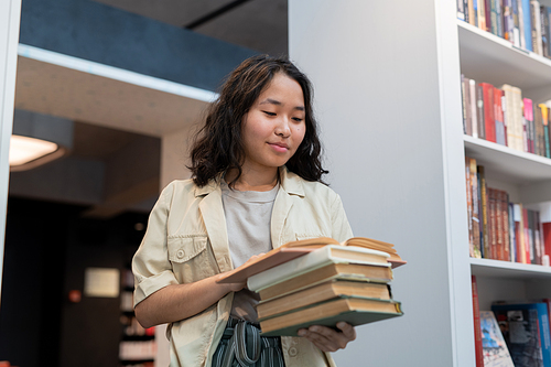 Happy girl with stack of books reading one of them in college library