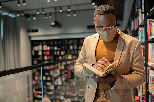 Young man of African ethnicity reading book while standing in large bookshop