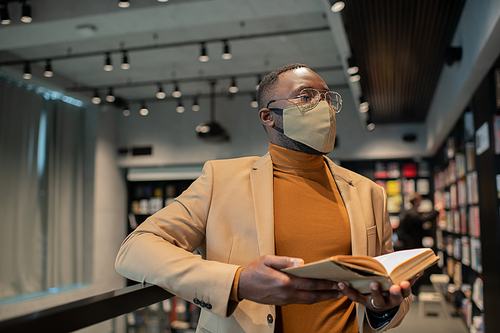 Young African man with open book looking at display with lots of literature in contemporary bookshop