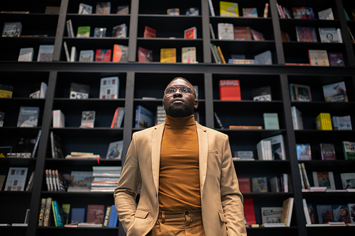 Young elegant businessman in suit and eyeglasses standing against large bookshelf in contemporary bookshop