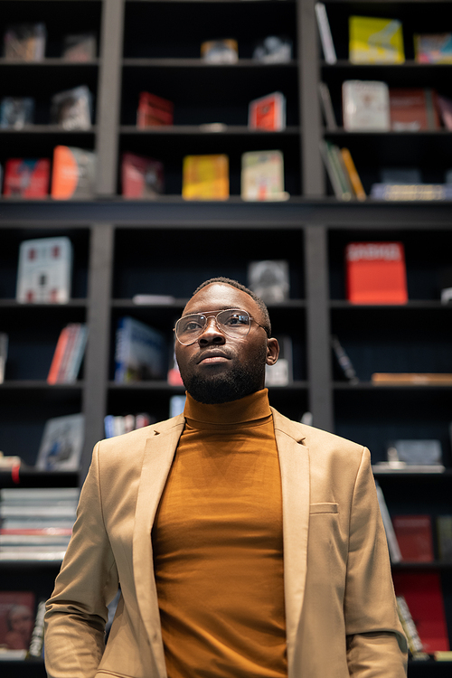 Young African-American man in smart casualwear standing in large bookshop against bookshelf