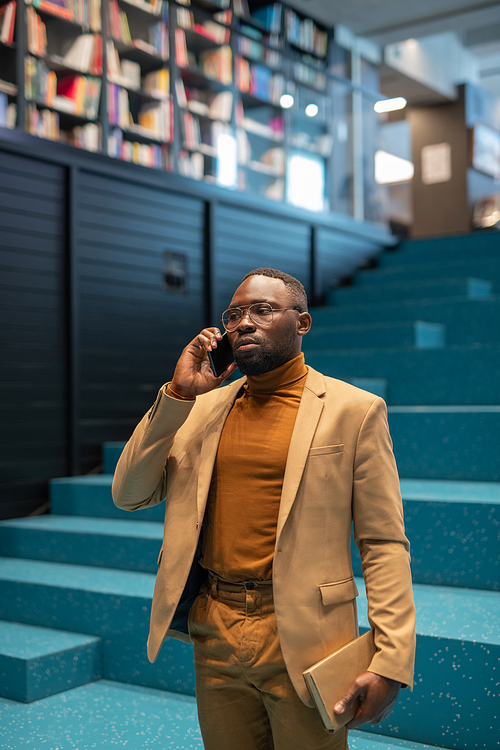 Young confident man with smartphone by ear and book in hand standing in large bookshop