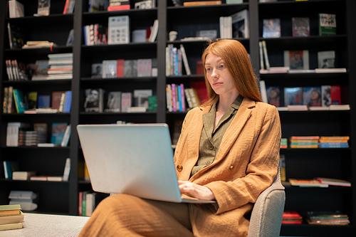 Young female with laptop looking through e-book on screen while sitting in bookshop