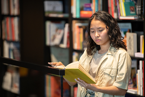 Young serious student reading book while standing by shelves with lots of books