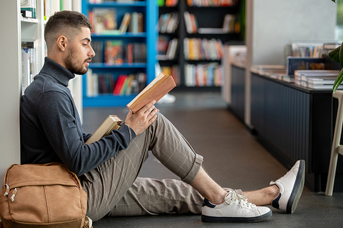 Serious young student in casualwear sitting against bookshelves and reading one of books