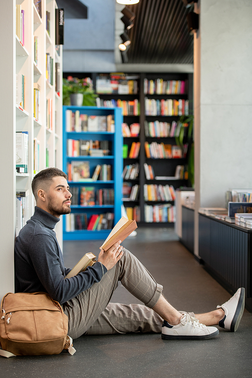 Young distracted man with open book looking aside while sitting on he floor against bookshelf