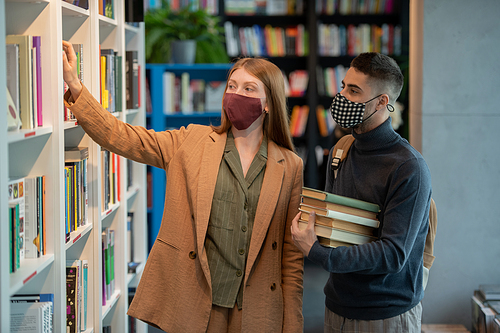 Small group of young students in protective masks choosing books in library