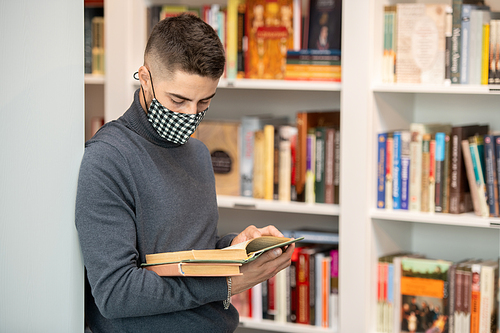 Young contemporary student in protective mask reading book while standing by bookshelves