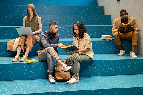 Intercultural couple of students discussing book while sitting on staircase against groupmates