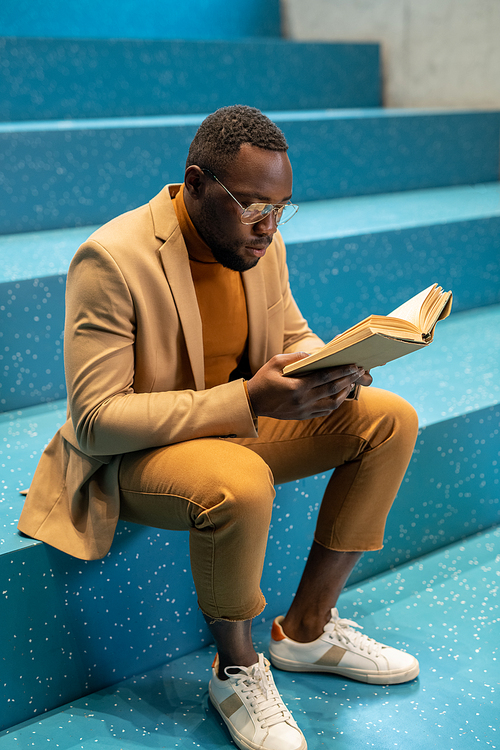 Contemporary African student reading book on blue staircase after classes