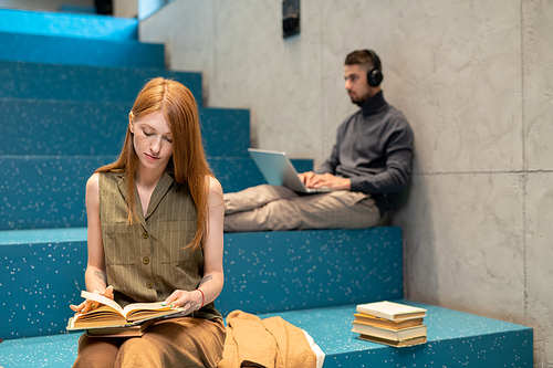 Young female with open book reading novel while sitting on step of blue staircase against guy with laptop