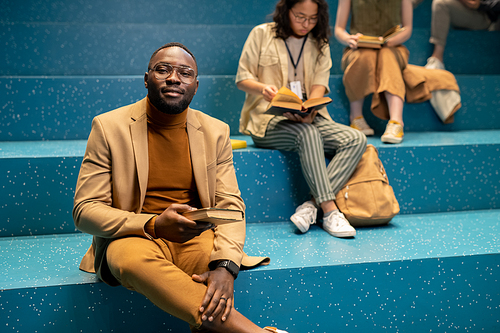 Happy African guy in smart casualwear holding book while sitting against two female students