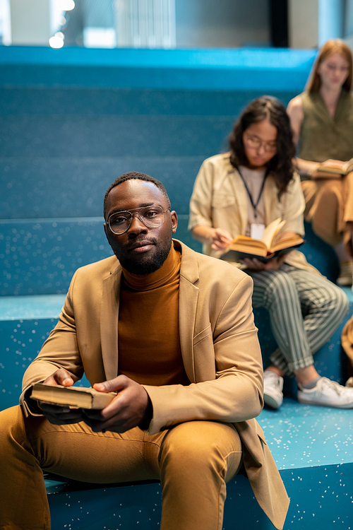 Young African businessman with book looking at you against his female colleagues